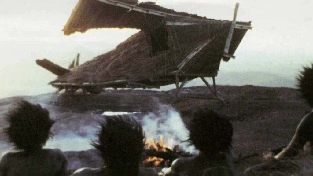 Photo of cargo cult people gathered around fire with straw airplane in background.